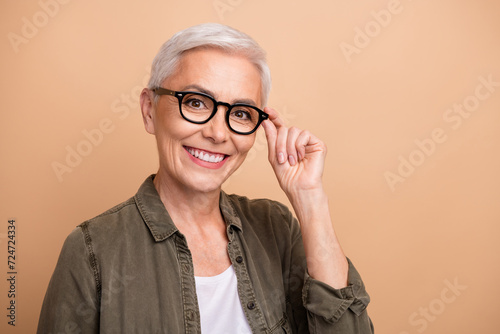 Portrait of satisfied business woman wearing khaki shirt touch eyeglases new style accessories advert isolated on beige color background photo