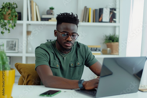Young African American man wearing jeans and green t-shirt sitting working in laptop in stylish white home office