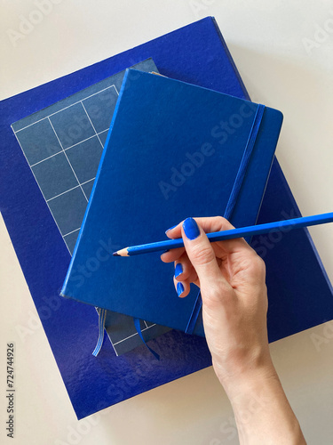Hand with blue nail polish, holding a blue pencil on a stack of blue books, notebooks and folders.