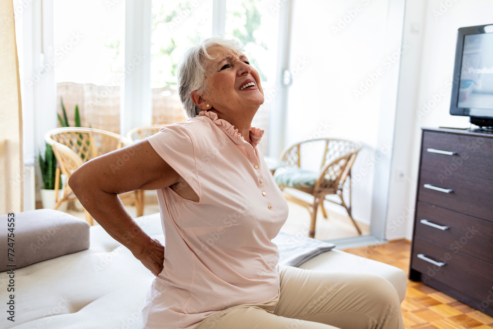 Unhappy mature woman touching back, feeling pain, sitting on couch in living room, unhealthy middle aged senior female suffering from backache, spinal problem, rubbing stiff muscles or kidney. Unhappy mature woman touching back, feeling pain, sitting on couch in living room, unhealthy middle aged senior female suffering from backache, spinal problem, rubbing stiff muscles or kidney.