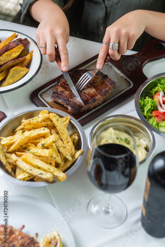 hands cutting meat in a table garnished by fries, salad and a cup of wine.