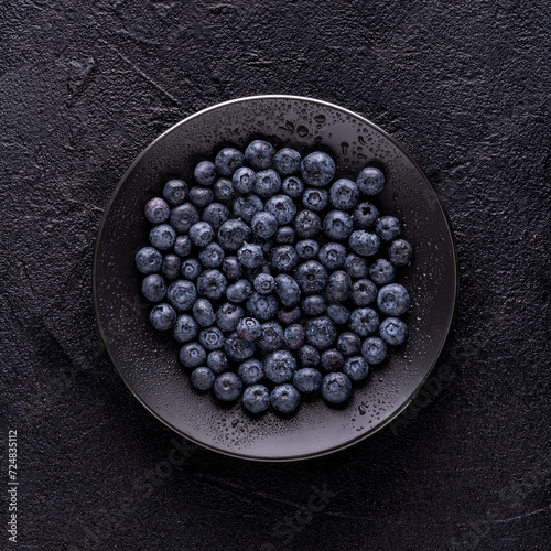 from above, on a black textured background, a black plate with fresh wet blueberries.