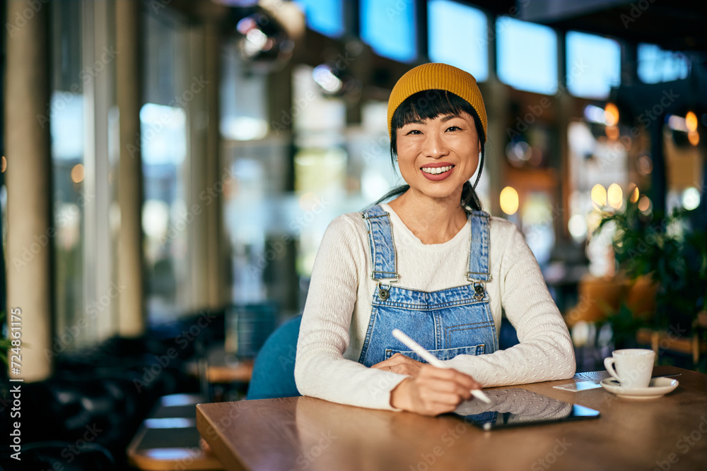 Portrait of a smiling Asian woman, using a digital tablet and a pen, making some designs online, working from the restaurant.