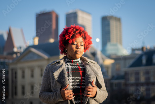 sexy moody female model with strong unique style appearance in urban inner city environment The Hague cityscape skyline. Confident black lady stylish red hair with winter good taste Holland fashion 
