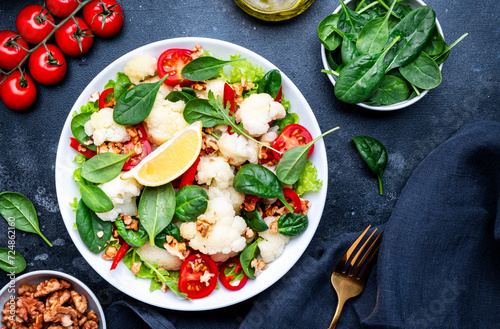 Healthy vegan salad with white cauliflower, baked red paprika, cherry tomatoes and spinach with walnuts, gray table background, top view