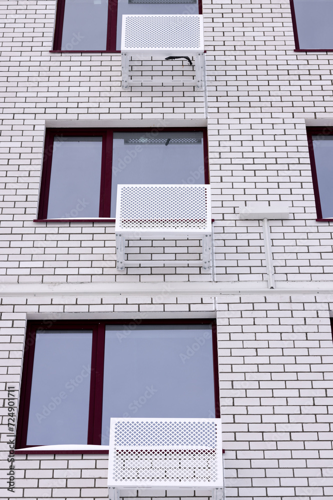 Metal white empty boxes for installing an air conditioner near the windows on the wall of a white brick house.