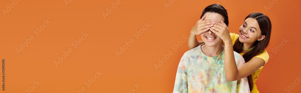 pretty teenage girl closing eyes of her asian friend on orange backdrop, friendship day, banner pretty teenage girl closing eyes of her asian friend on orange backdrop, friendship day, banner