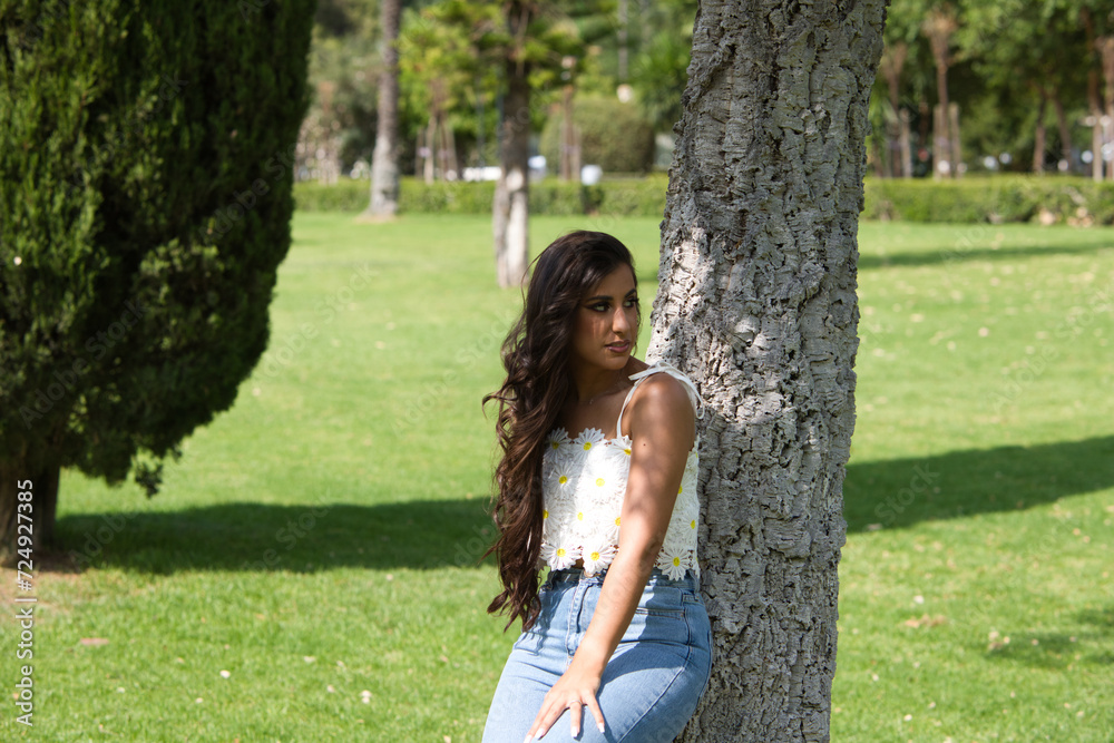 Beautiful Spanish brunette woman with long curly hair and big slit eyes leans her back against the trunk of a tree. The woman is smiling and happy and enjoying the day in the city of Seville in Spain. Beautiful Spanish brunette woman with long curly hair and big slit eyes leans her back against the trunk of a tree. The woman is smiling and happy and enjoying the day in the city of Seville in Spain.