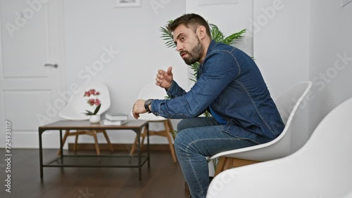 Stressed out young hispanic man, desperately checking time, sitting anxiously on chair in waiting room, embodying the problem of pressure