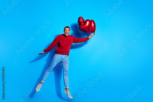 Full body portrait of cheerful nice young man arm hold heart figure balloons jump empty space isolated on blue color background