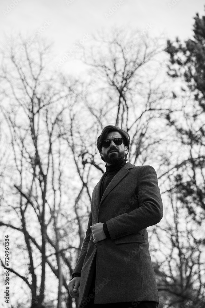 Outside image of a young bearded man in a coat and sunglasses poses alone, tree background. Outside image of a young bearded man in a coat and sunglasses poses alone, tree background.