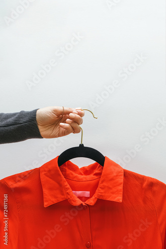 Female hand holding a hanger with a red feminine shirt white wall.