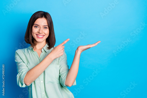 Portrait of cheerful girl with stylish hair wear teal shirt directing at offer on arm empty space isolated on blue color background