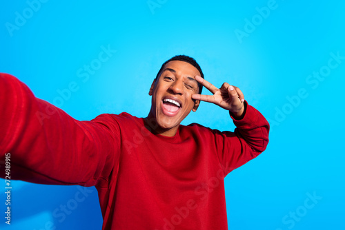 Photo of funky cheerful guy take selfie demonstrate v-sign eye wink blink isolated on blue color background