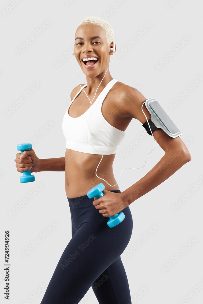 smiling woman doing fitness workout, lifts dumbbells while listening music through earphones of mobile phone on arm band. Latin American female athlete. Sportswoman do training, isolated on background smiling woman doing fitness workout, lifts dumbbells while listening music through earphones of mobile phone on arm band. Latin American female athlete. Sportswoman do training, isolated on background