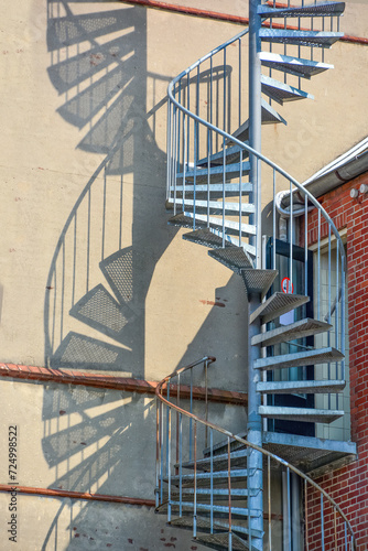Vertical view of a blue circulair steel fire escape on the outside of a building with shadows of the steps and railing on the wall photo