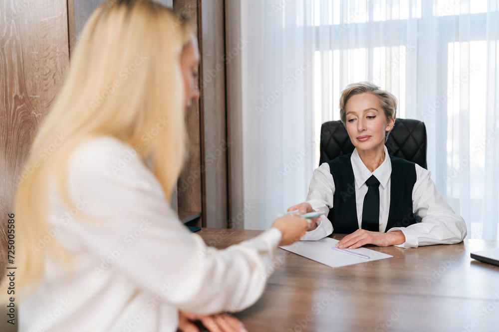 Rear view of unrecognizable female investor giving euro banknotes to middle-aged advisor sitting at office desk. Young woman talking to real estate agent, planning future. Concept of finance. Rear view of unrecognizable female investor giving euro banknotes to middle-aged advisor sitting at office desk. Young woman talking to real estate agent, planning future. Concept of finance.