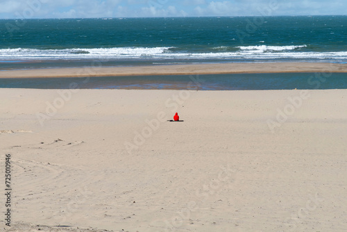 Panoramic view over an empty beach other than one person in red shirt sitting in the middle overlooking the ocean and far away horizon with waves crashing on the beach