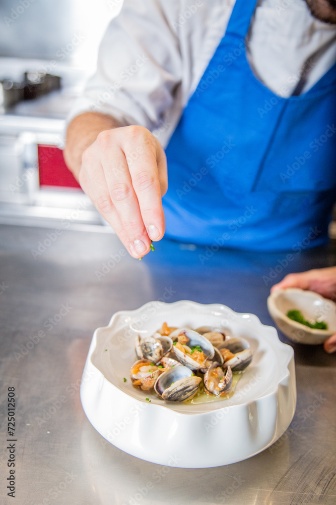 Chef decora plato de almejas a la marinera con cebollino y salsa de vino blanco albariño sobre un plato blanco en la barra de un restaurante Chef decora plato de almejas a la marinera con cebollino y salsa de vino blanco albariño sobre un plato blanco en la barra de un restaurante