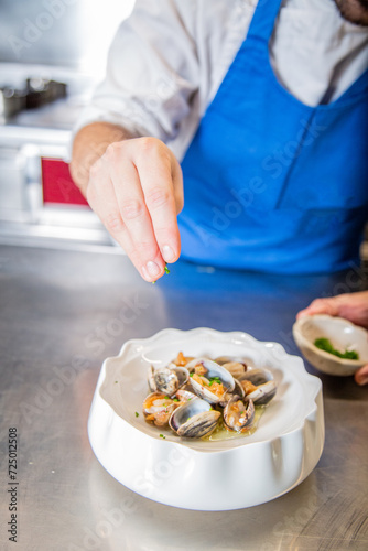 Chef decora plato de almejas a la marinera con cebollino y salsa de vino blanco albariño sobre un plato blanco en la barra de un restaurante