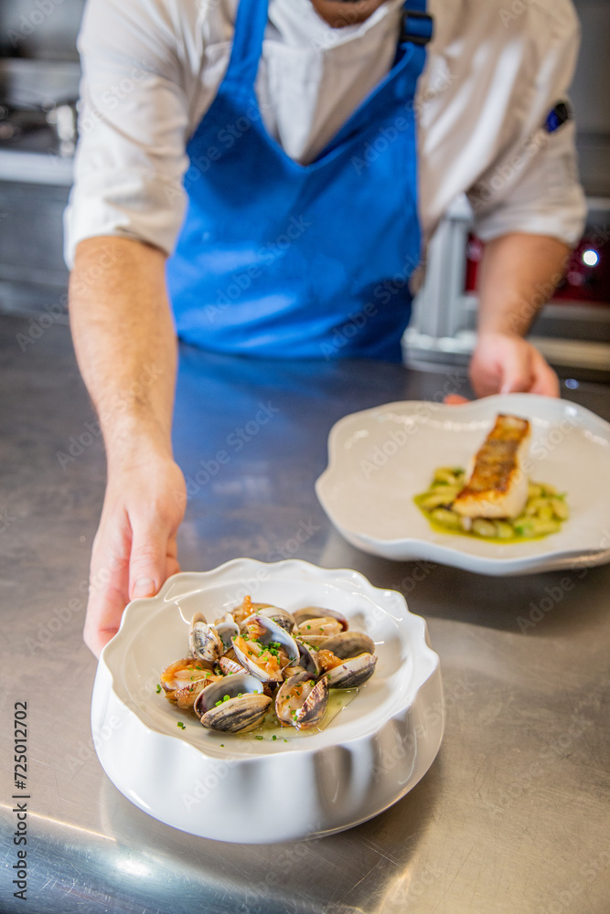 Chef decora plato de almejas a la marinera con cebollino y salsa de vino blanco albariño sobre un plato blanco en la barra de un restaurante Chef decora plato de almejas a la marinera con cebollino y salsa de vino blanco albariño sobre un plato blanco en la barra de un restaurante