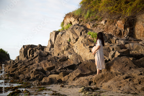 Mujer joven preciosa embarazada pasea y camina por la playay las rocas cerca del mar en un atardecer en la costa