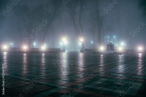 The alley of a night winter park in a fog. Footpath in a fabulous winter city park at night in fog with benches and latterns. Beautiful foggy evening in the Mariinsky Park. Kyiv, Ukraine.