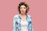Portrait of funny childish attractive woman with curly hairstyle wearing blue shirt standing looking at camera showing tongue out. Indoor studio shot isolated on pink background.