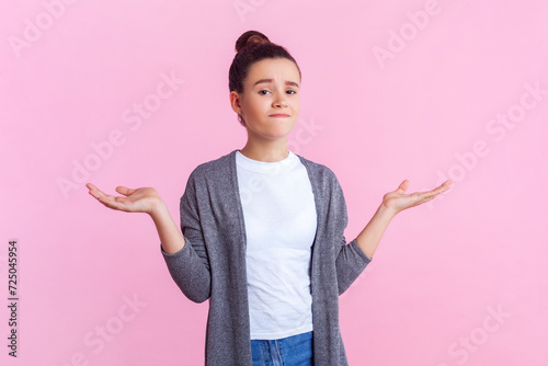Portrait of confused teenage girl with bun hairstyle in casual clothes standing with shrugging shoulders having uncertain facial expression. Indoor studio shot isolated on pink background.