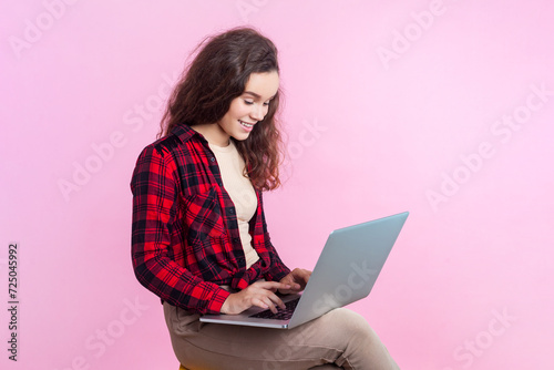 Portrait of smiling teenage girl freelancer with wavy hair in red checkered shirt sitting typing on laptop keyboard writing article. Indoor studio shot isolated on pink background.