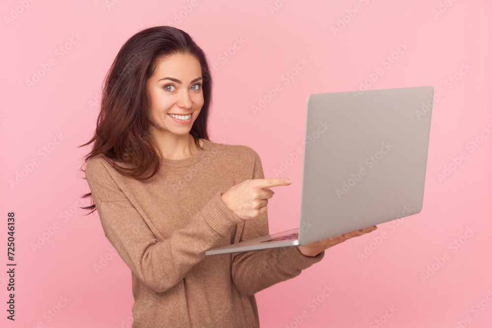 Side view portrait of beautiful woman with wavy hair, holding laptop pointing at display having online work wearing wearing brown pullover. Indoor studio shot isolated on pink background Side view portrait of beautiful woman with wavy hair, holding laptop pointing at display having online work wearing wearing brown pullover. Indoor studio shot isolated on pink background
