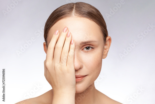 Closeup portrait of calm shy beautiful woman with perfect skin covering half of face with palm lookingat camera. Indoor studio shot isolated over gray background. photo