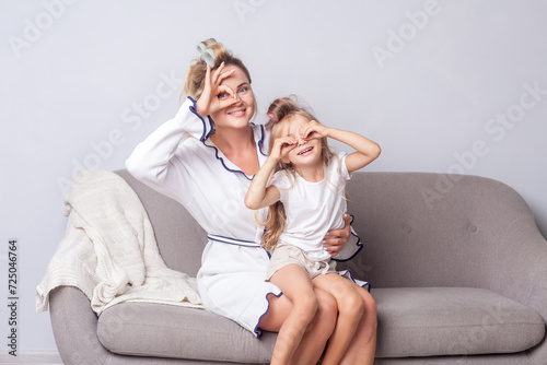 Happy woman and daughter looking at camera through fingers in binoculars gesture and smiling having fun together at home sitting on sofa, expressing positive emotions.