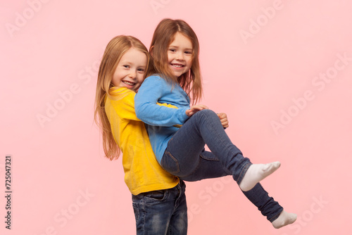 Portrait of two playful cute positive little girls having fun together, playing active games, expressing joy and happiness. Indoor studio shot isolated on pink background.
