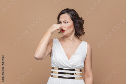 Confused unhappy middle aged woman with wavy hair holding breath with fingers on nose, turning away from stinky odor, fart, wearing white dress. Indoor studio shot isolated on light brown background.
