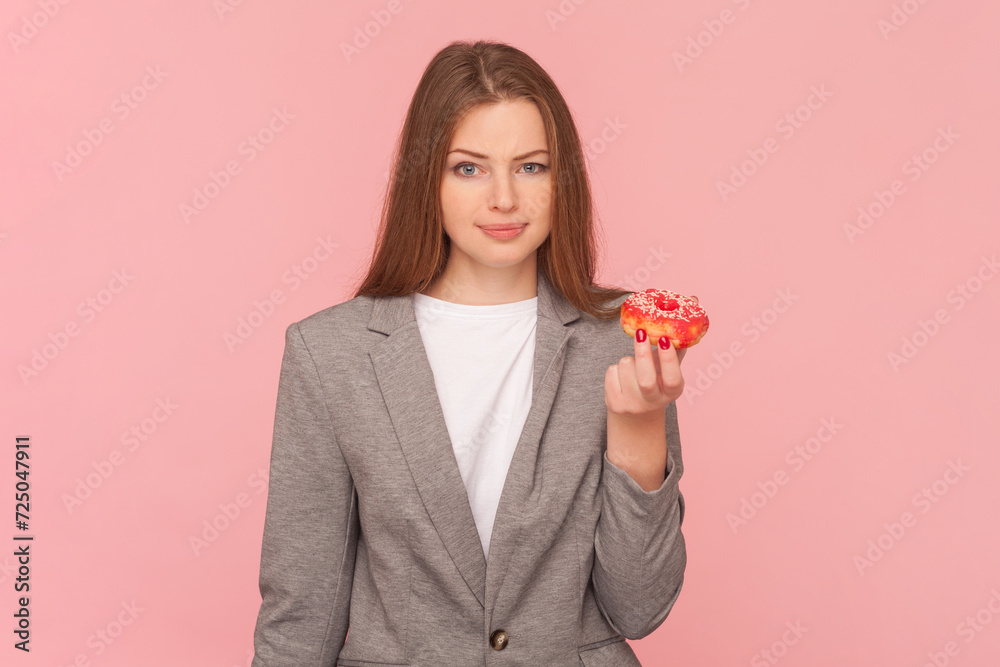 Portrait of confused doubtful woman standing with sugary dessert in hands, looking at camera, deciding eating or not, wearing business suit. Indoor studio shot isolated on pink background.