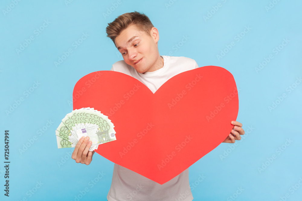 Portrait of rich delighted man wearing white t-shirt holding big red heart and fan of euro banknotes, love to money. Indoor studio shot isolated on blue background Portrait of rich delighted man wearing white t-shirt holding big red heart and fan of euro banknotes, love to money. Indoor studio shot isolated on blue background