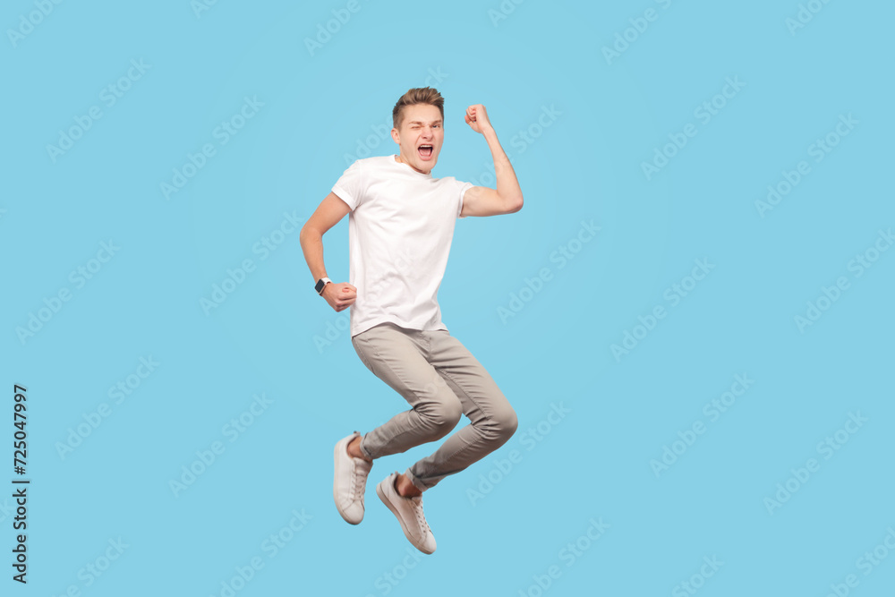 Full length portrait of satisfied joyful positive man wearing white t-shirt jumping with clenched fists, celebrating his victory. Indoor studio shot isolated on blue background
