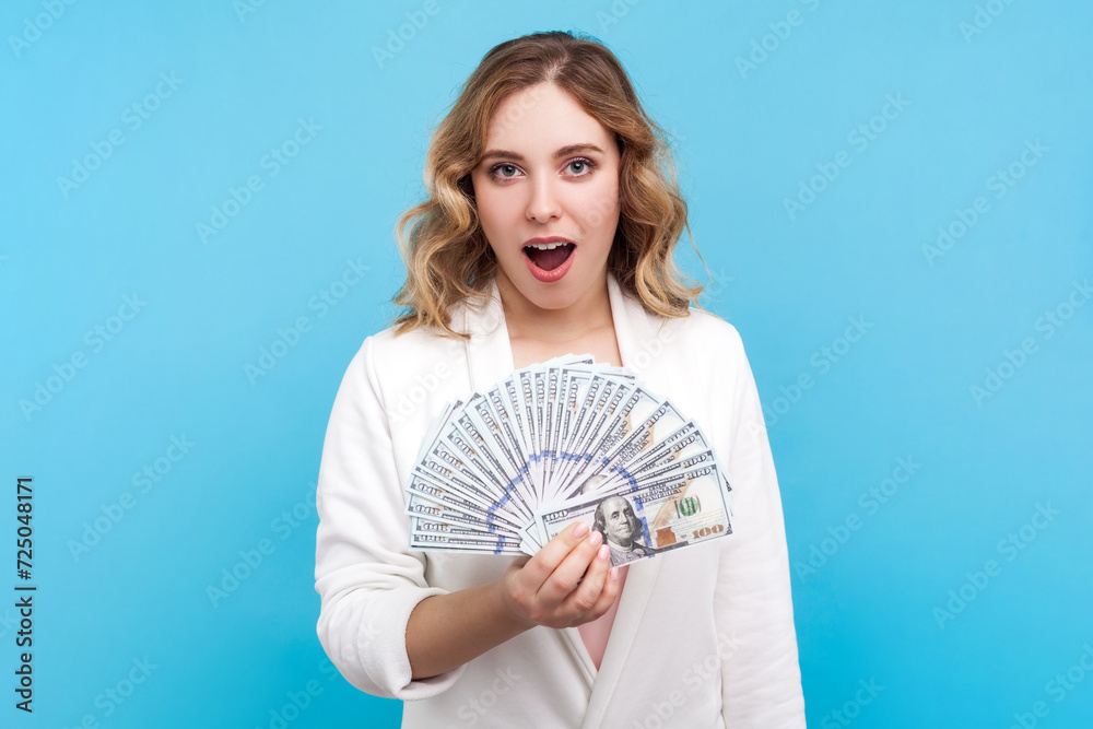 Portrait of amazed shocked blond woman with wavy hair standing with fan of money saying wow earning big sum of money, wearing white shirt. Indoor studio shot isolated on blue background. Portrait of amazed shocked blond woman with wavy hair standing with fan of money saying wow earning big sum of money, wearing white shirt. Indoor studio shot isolated on blue background.