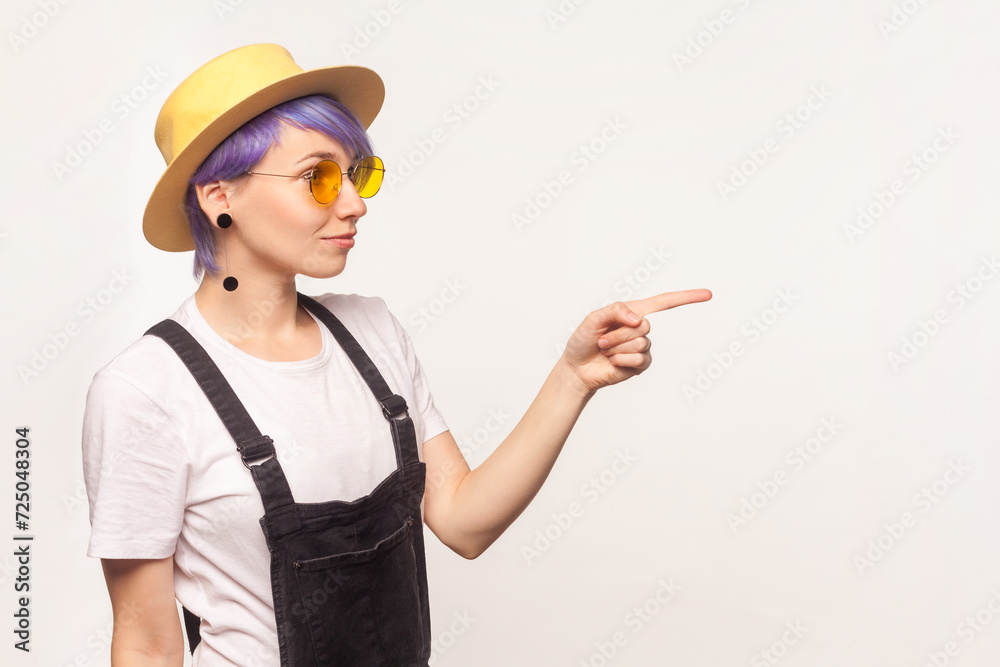 Side view portrait of hipster woman with violet hair in sunglasses and hat pointing at copy space for advertisement, empty area. Indoor studio shot isolated on white background. Side view portrait of hipster woman with violet hair in sunglasses and hat pointing at copy space for advertisement, empty area. Indoor studio shot isolated on white background.
