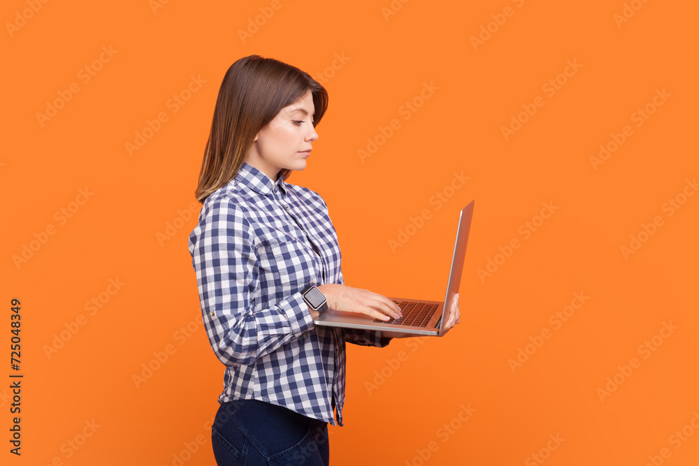 Side view portrait of confident serious woman with brown hair working on notebook, online project, work from home, wearing checkered shirt. Indoor studio shot isolated on orange background Side view portrait of confident serious woman with brown hair working on notebook, online project, work from home, wearing checkered shirt. Indoor studio shot isolated on orange background