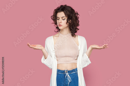 Portrait of doubtful pensive woman with curly hair wearing casual style outfit spreading palms aside, showing copy space, making decision. Indoor studio shot isolated on pink background.