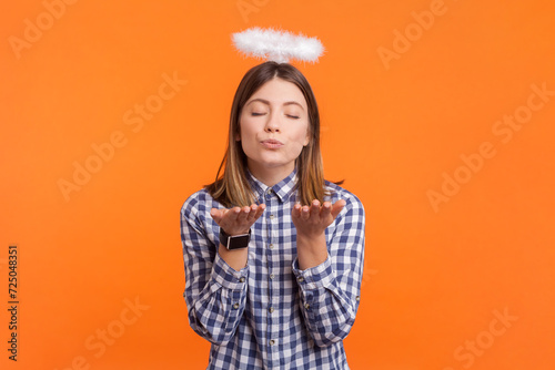 Portrait of romantic angelic woman with brown hair and nimb over head closed her eyes sending air kissing, wearing checkered shirt. Indoor studio shot isolated on orange background