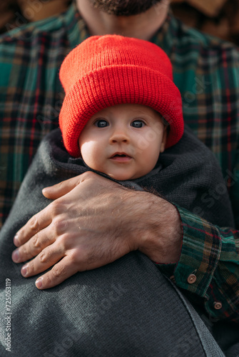 Portrait of a small child in a red hat. A small child in the arms of his father. A beautiful portrait of a child in his arms. Beautiful baby photo. Portrait of a small child. Dad hugs the baby © MISHA