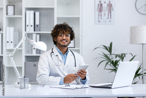 Cheerful young male healthcare professional with stethoscope sitting at his desk in a medical office, working on laptop and holding a tablet.