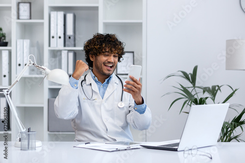 A joyful young healthcare professional in a clinic office rejoices while looking at his smartphone, expressing success or good news.