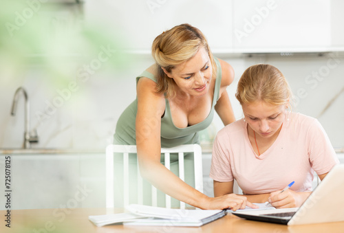 Teenage girl student engrossed in studies, sitting at table with laptop and diligently writing notes in notebook guided by positive friendly female tutor