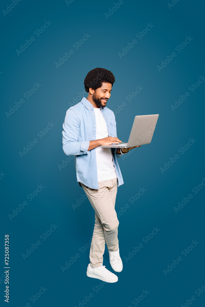 A young Brazilian man, male freelancer or student, standing and using a laptop, a picture of modern productivity on blue backdrop, representing the flexibility of remote work and digital nomadism
