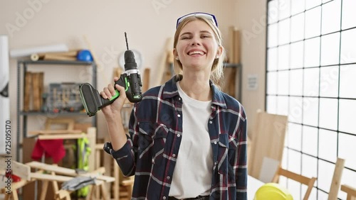 A smiling woman holding a drill in a bright carpentry workshop, portraying skill and confidence.