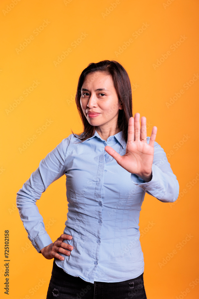 Serious asian woman raising palm to advertise stop gesture in studio over yellow background, expressing denial and refusal. Negative young adult showing rejection gesture, doing rejection symbol Serious asian woman raising palm to advertise stop gesture in studio over yellow background, expressing denial and refusal. Negative young adult showing rejection gesture, doing rejection symbol